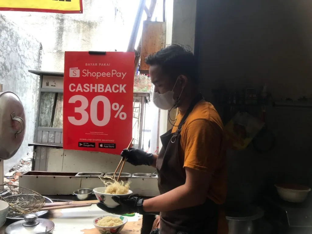 Seorang pedagang bakmi sedang sibuk meracik bumbu dan mie di dalam mangkuk di sebuah kedai bakmi di Bandung, dengan kepulan asap tipis dari panci rebusan.